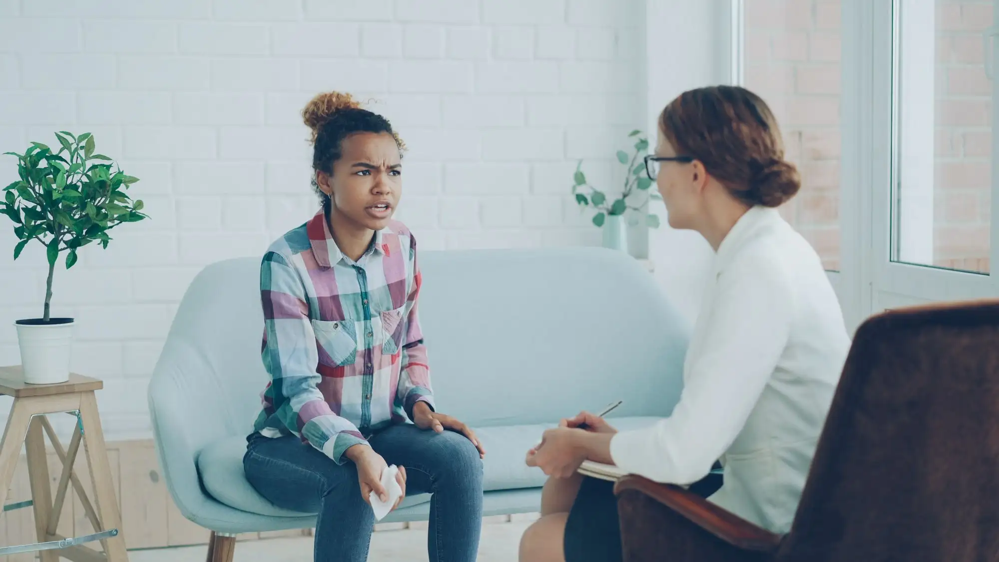 A counselor listens to a young woman on a sofa in a bright room.