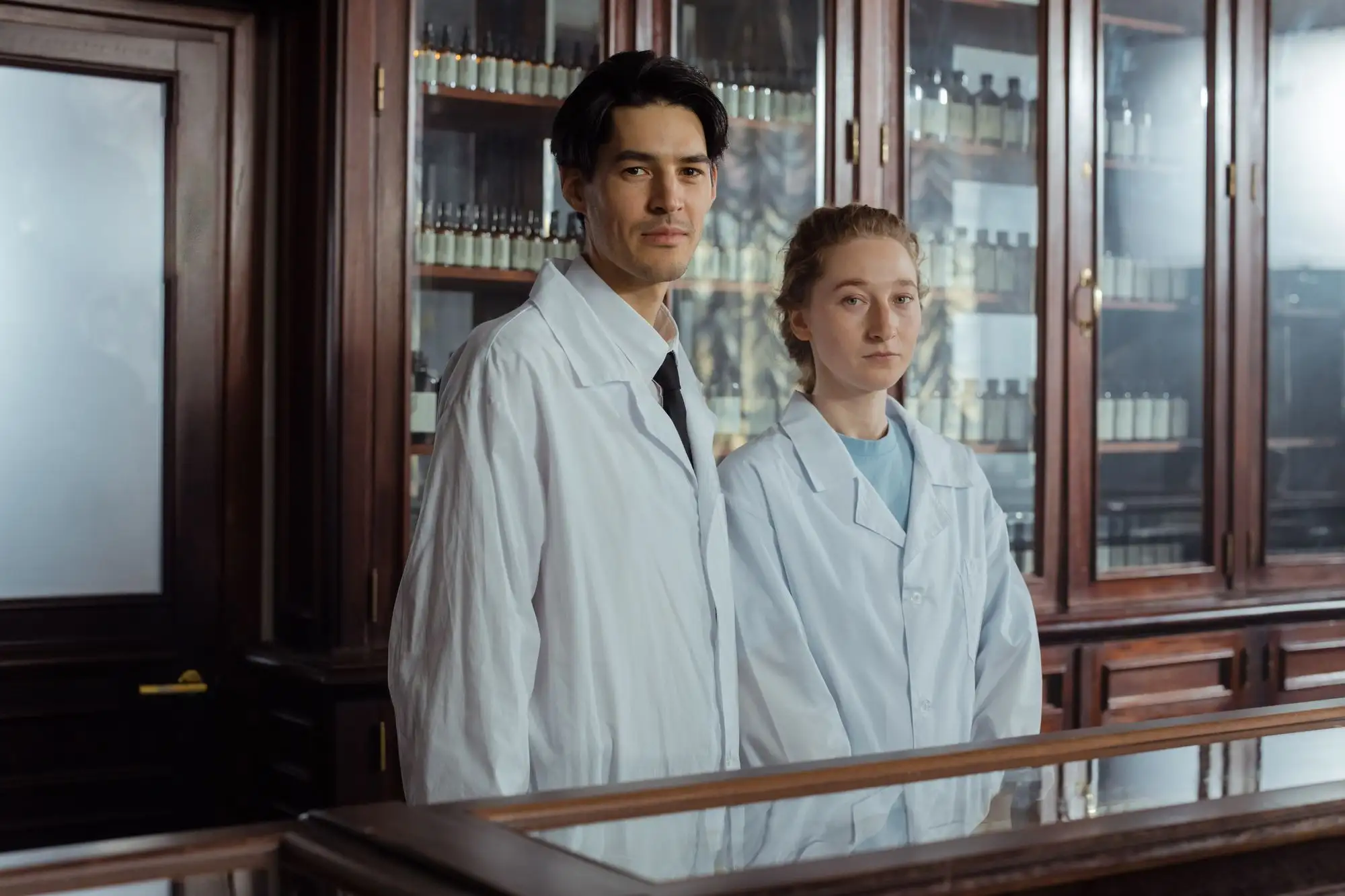 Two pharmacists in white coats stand behind a dispensary counter.
