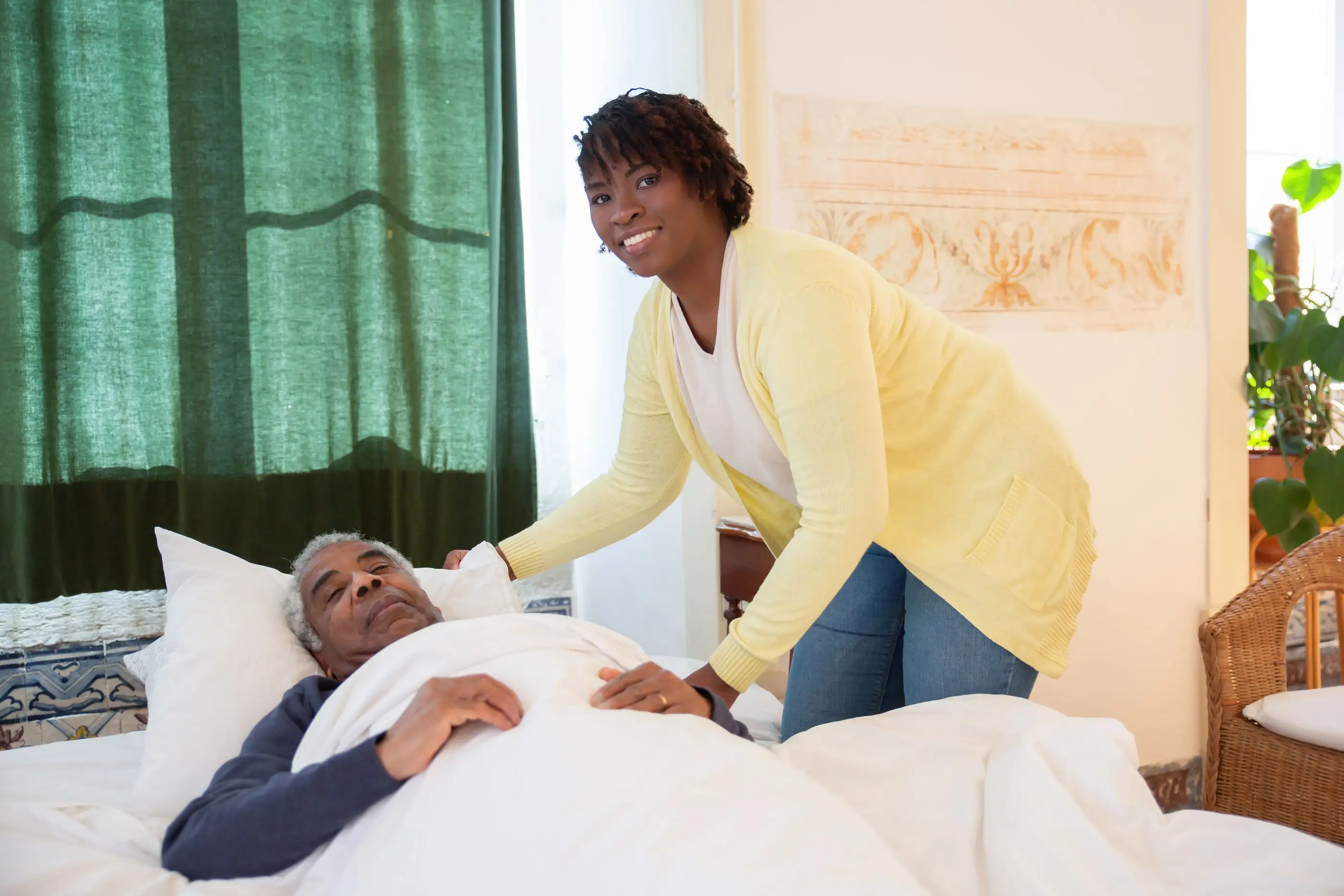 A caregiver smiles as she tucks in an older man resting in bed.