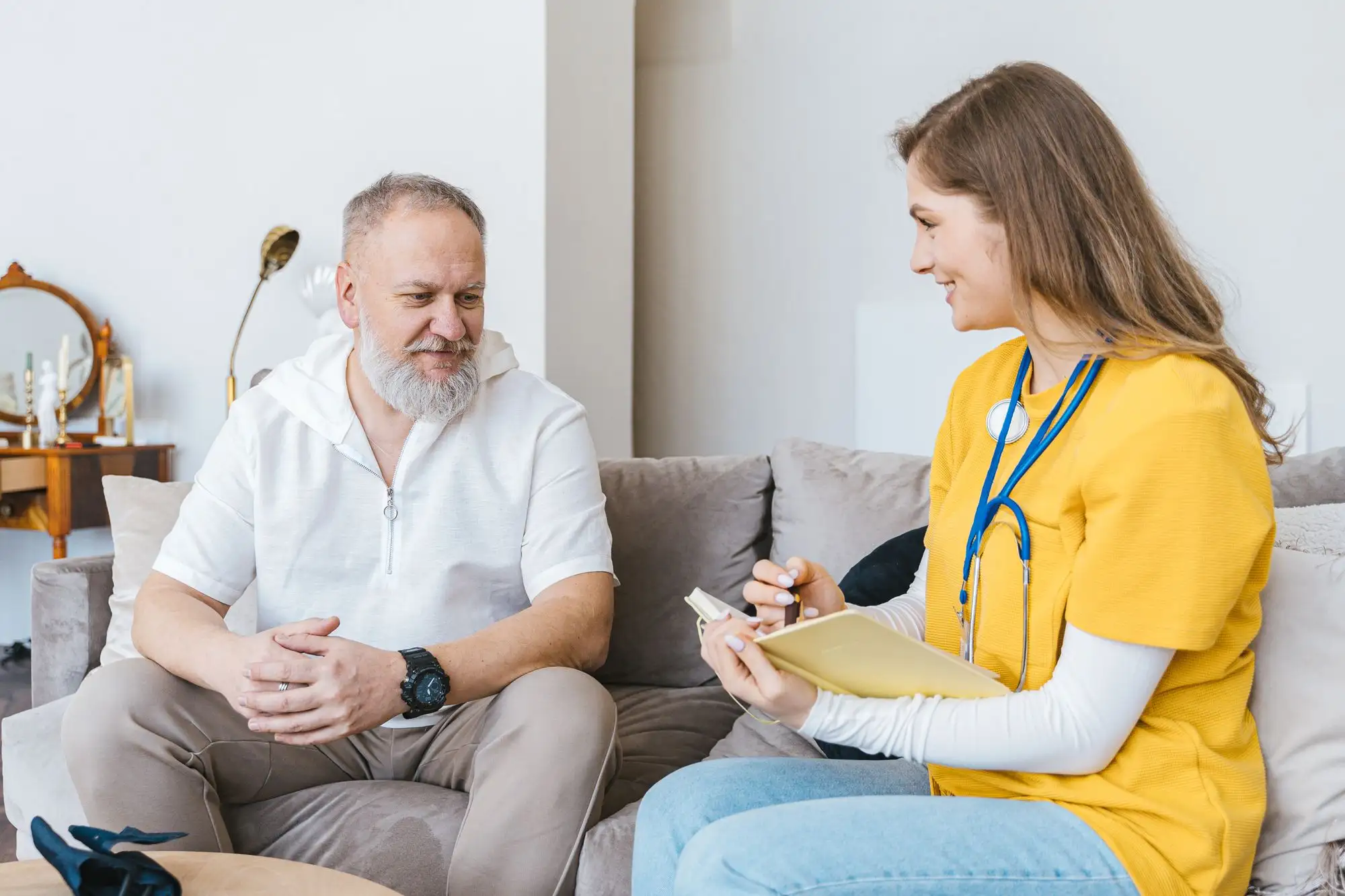 A clinician in a yellow uniform takes notes while talking with an older man at home.