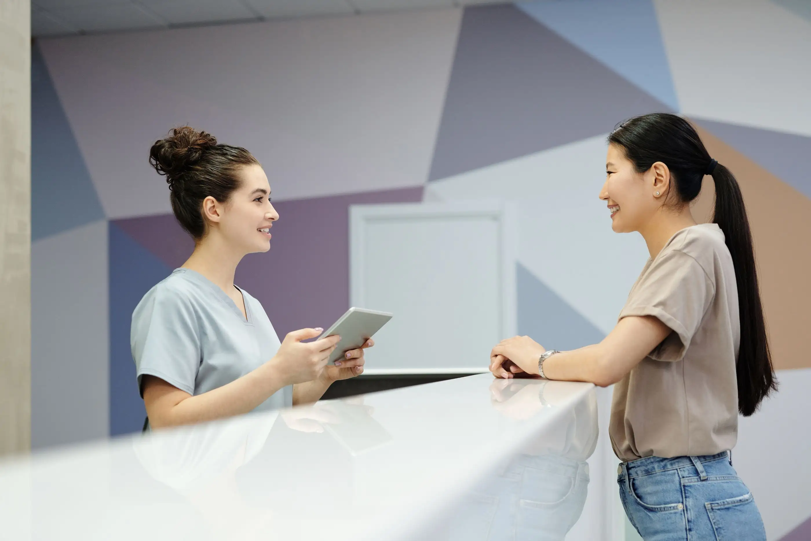 A clinic receptionist shows a patient information on a tablet across the desk.