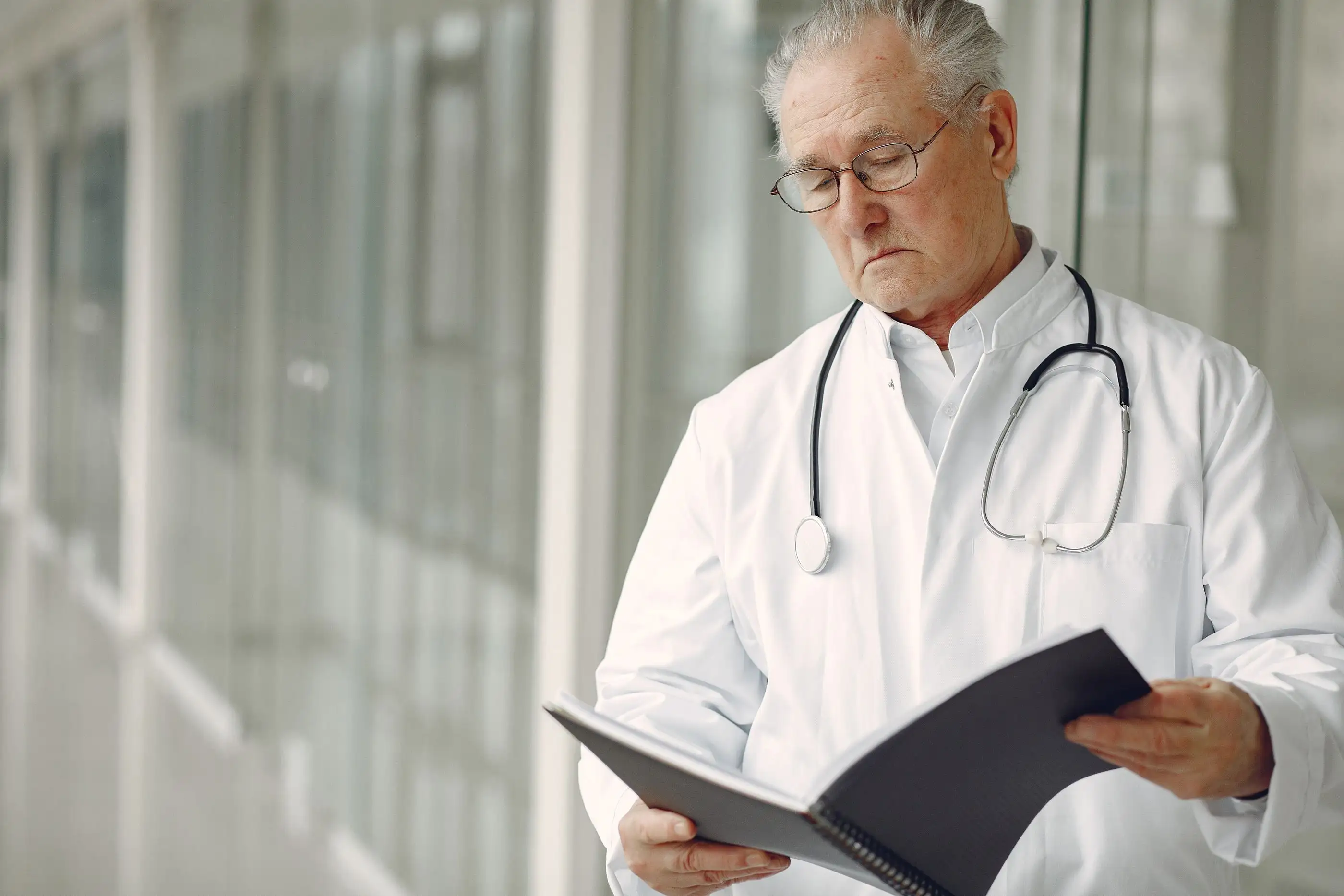 An older physician reads from a folder by a bright window.