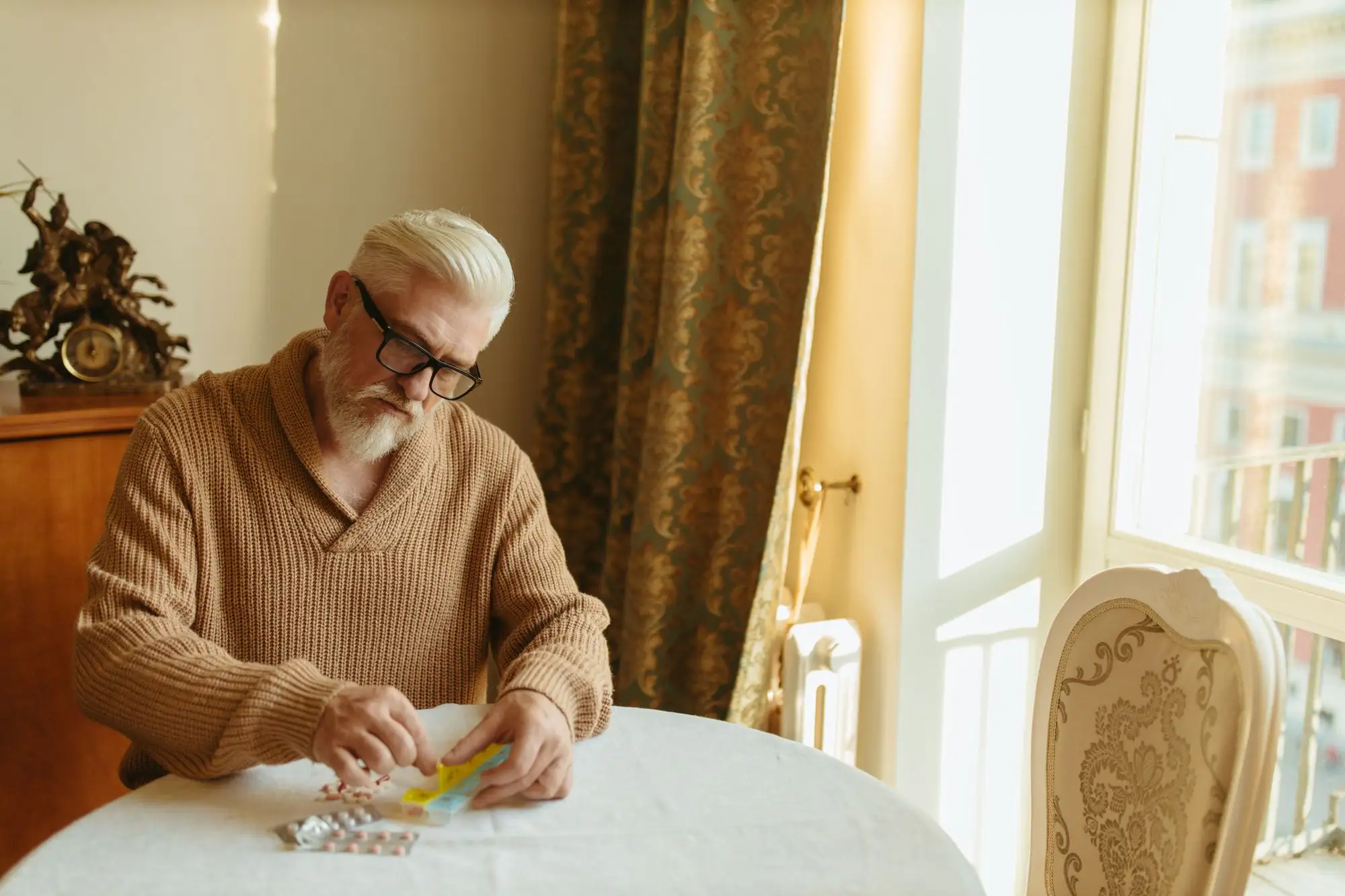 An older man sorts tablets into an organizer at a sunny table.