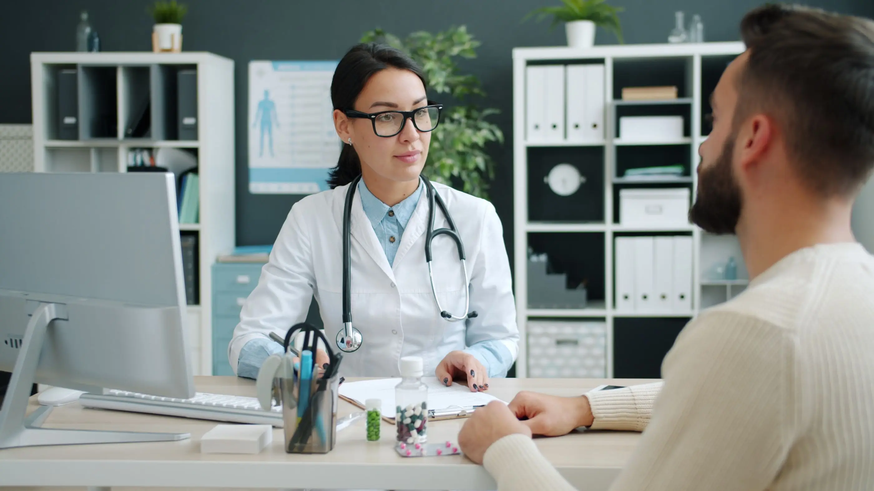 A clinician in a white coat listens to a patient across her desk.