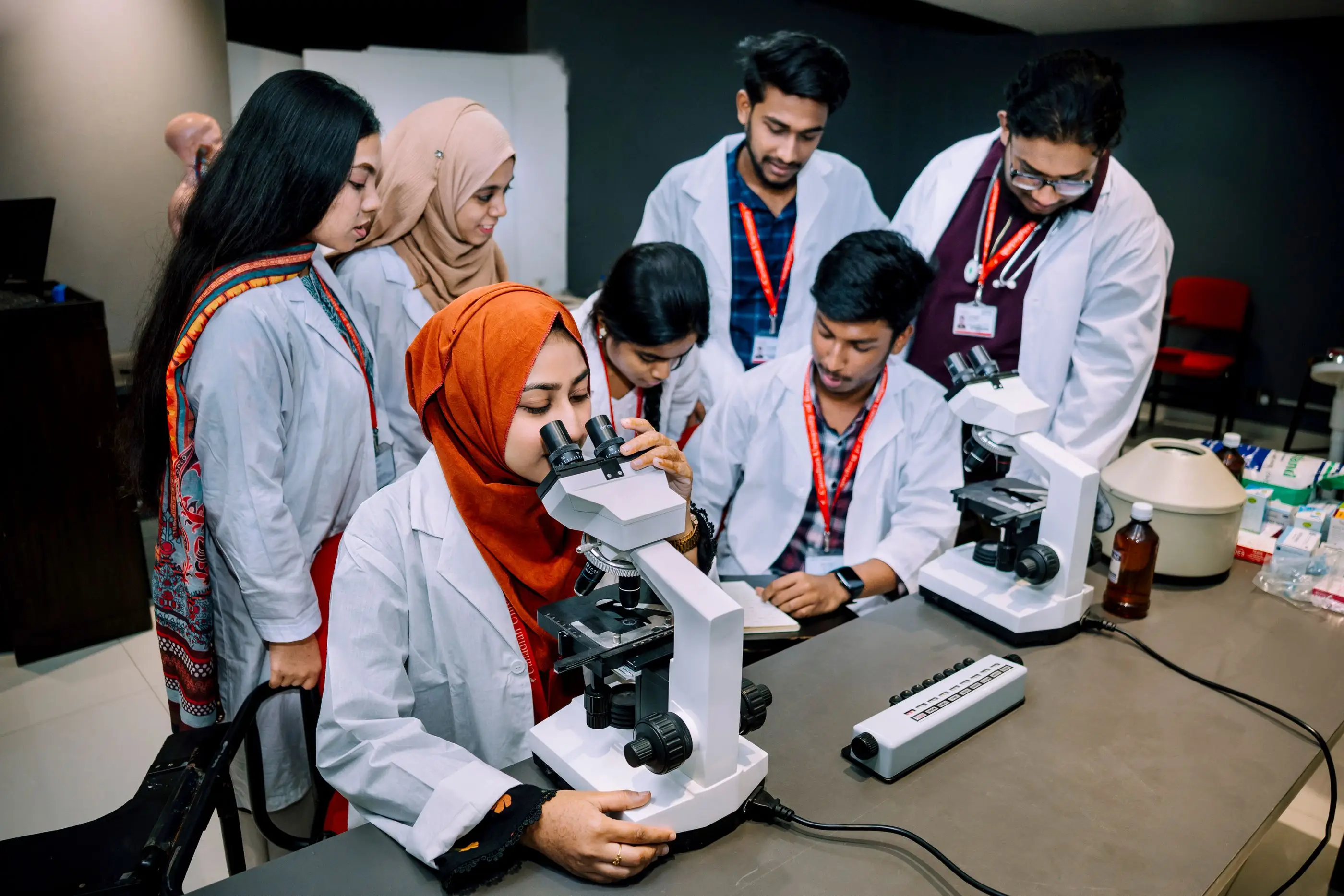 Pharmacy students in white coats work with microscopes in a teaching lab.