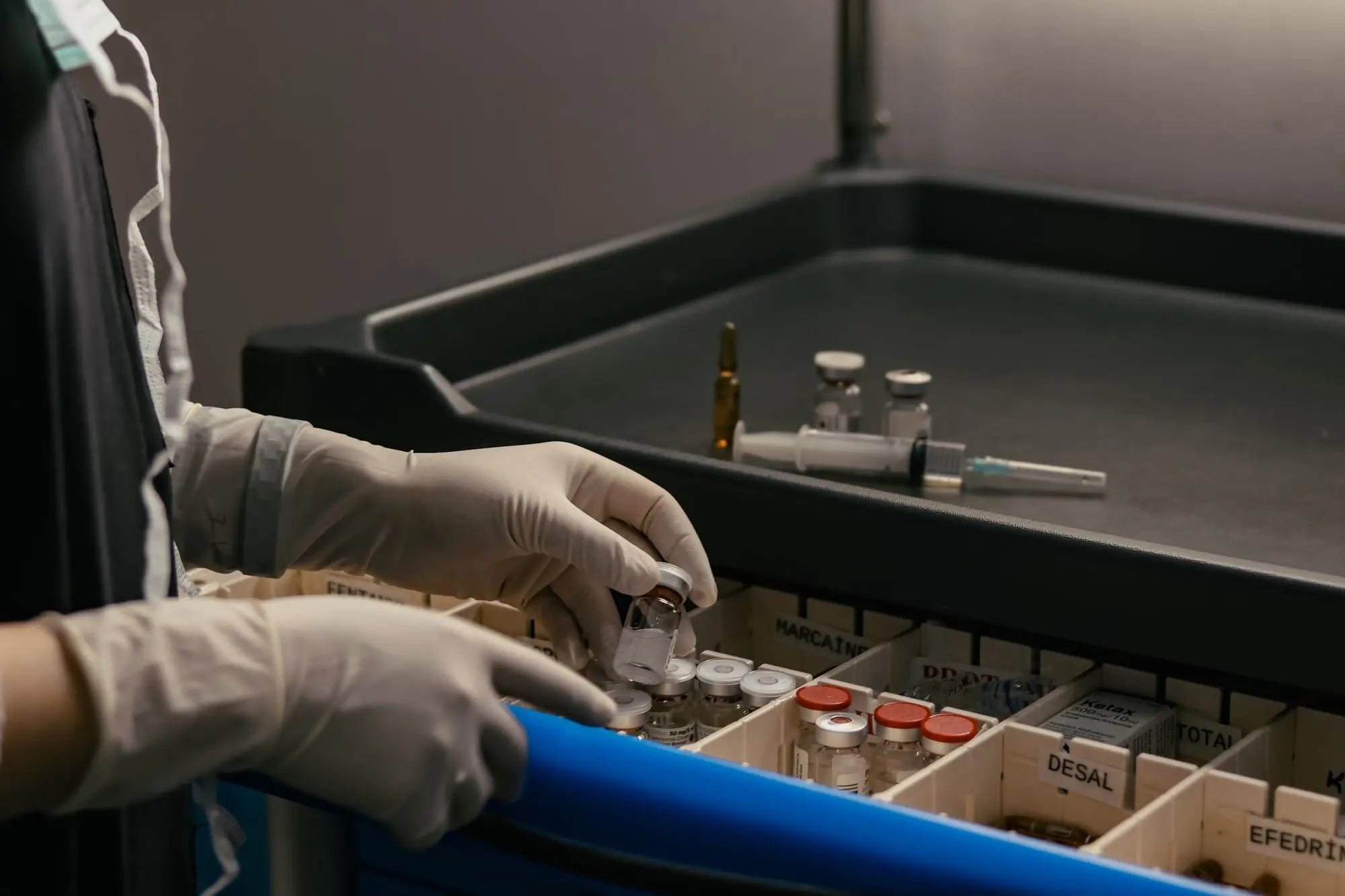 Gloved hands take a vial from a medication cart drawer.