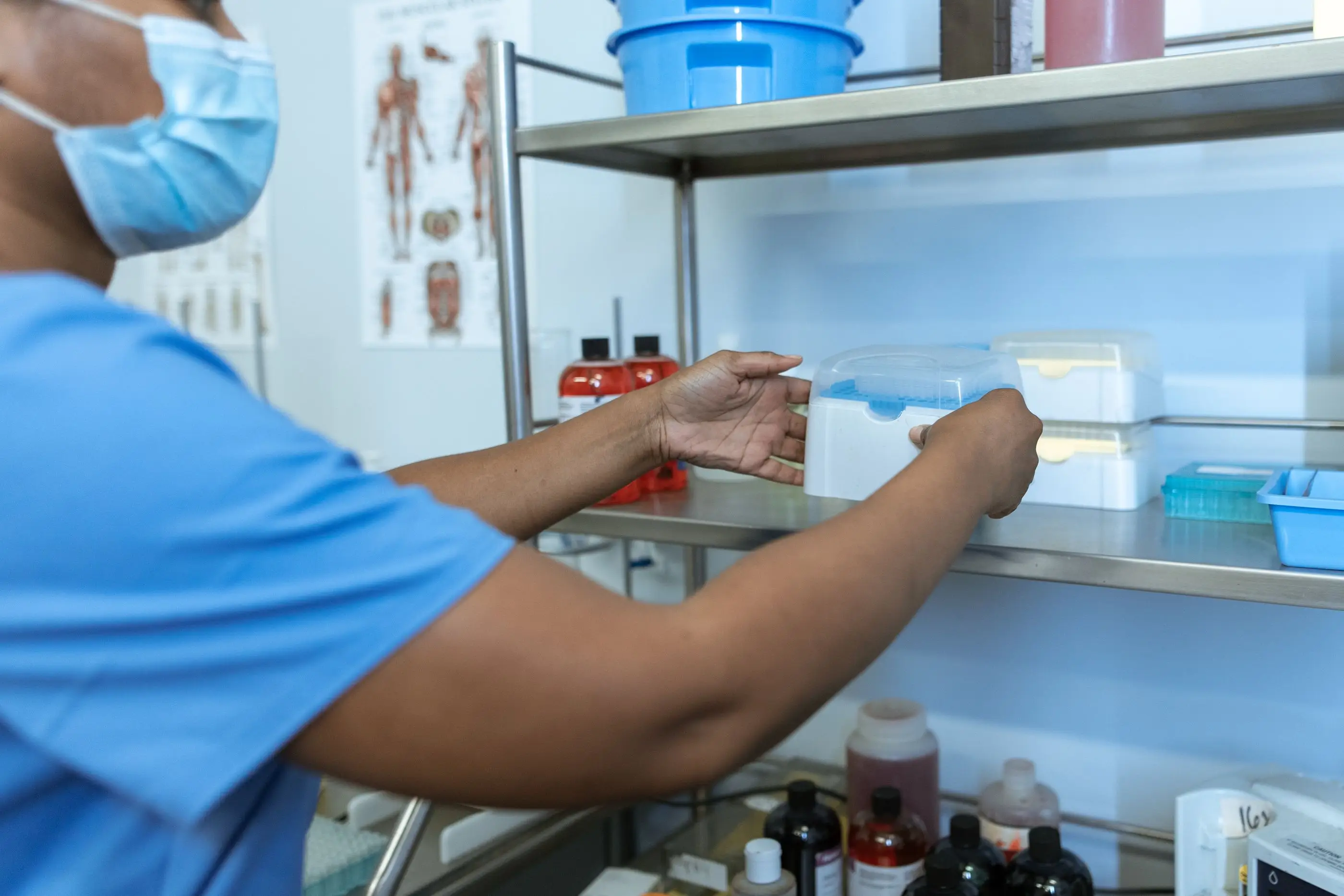A technician in scrubs takes supplies from pharmacy shelving.