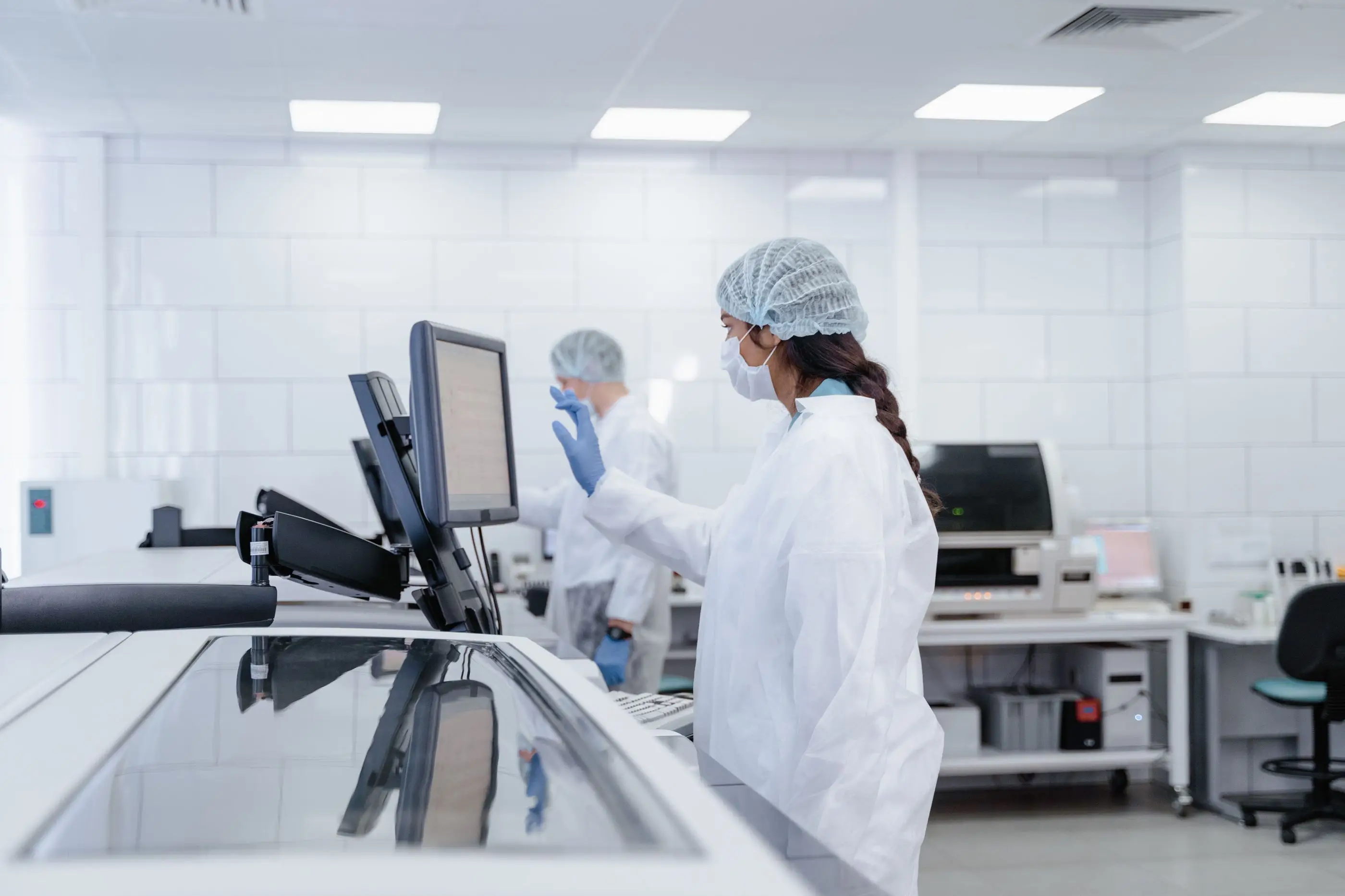 A technician in a lab coat and hair cover works in a bright laboratory.