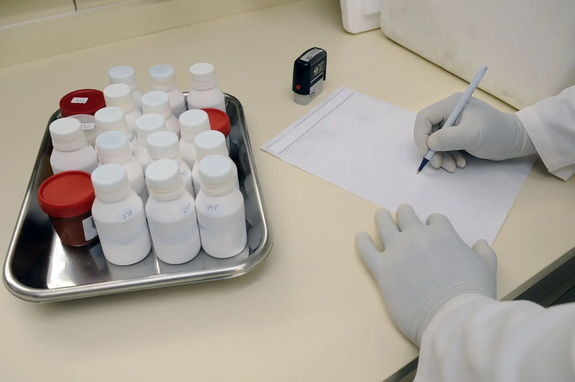 A gloved hand records a tray of labeled medication bottles.