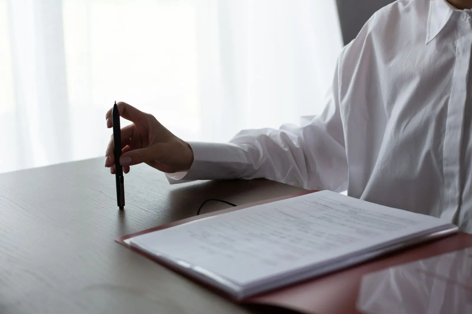 A hand holds a pen over a printed document in a folder on a desk.
