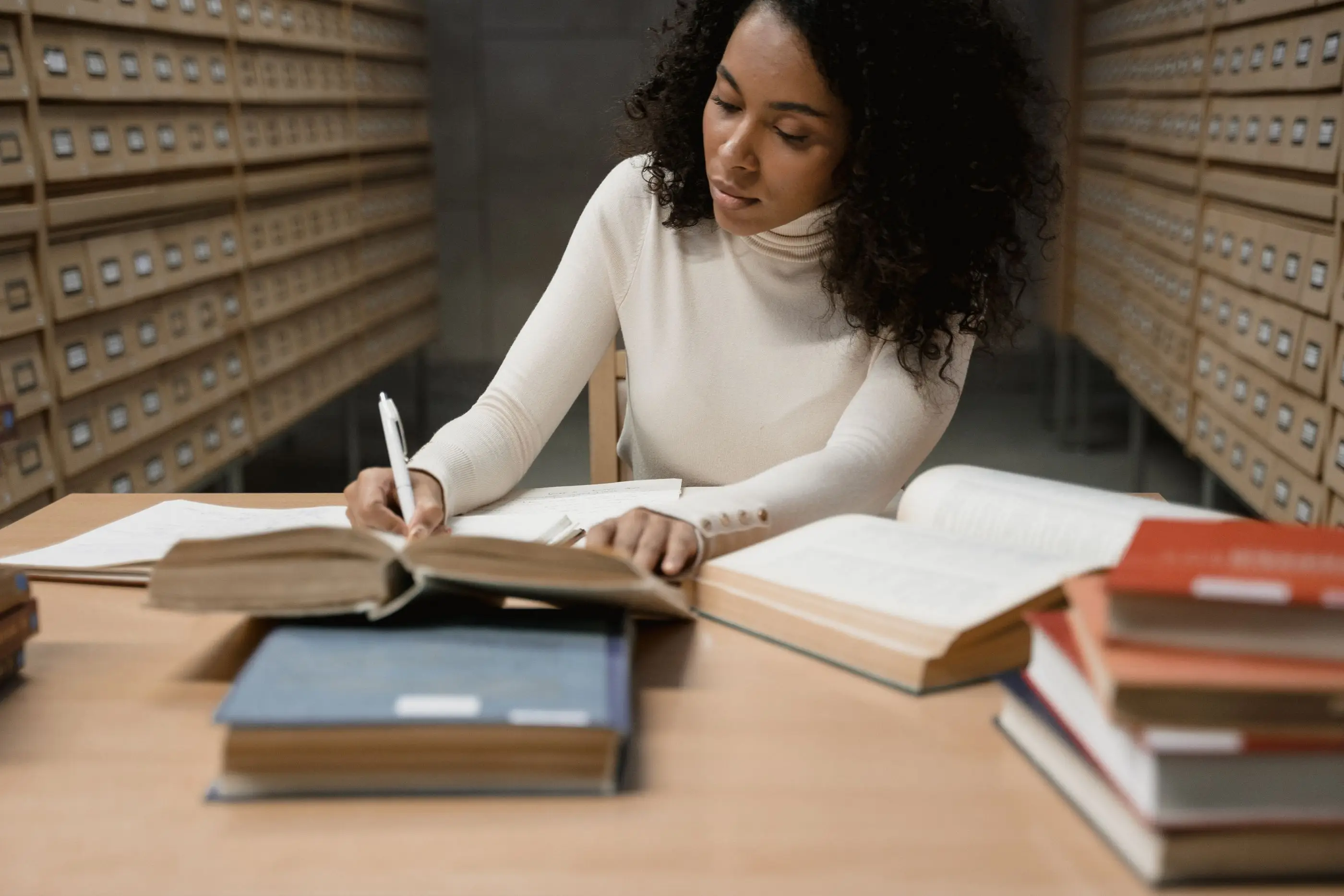 A woman takes notes from open reference books in front of a library card catalog.