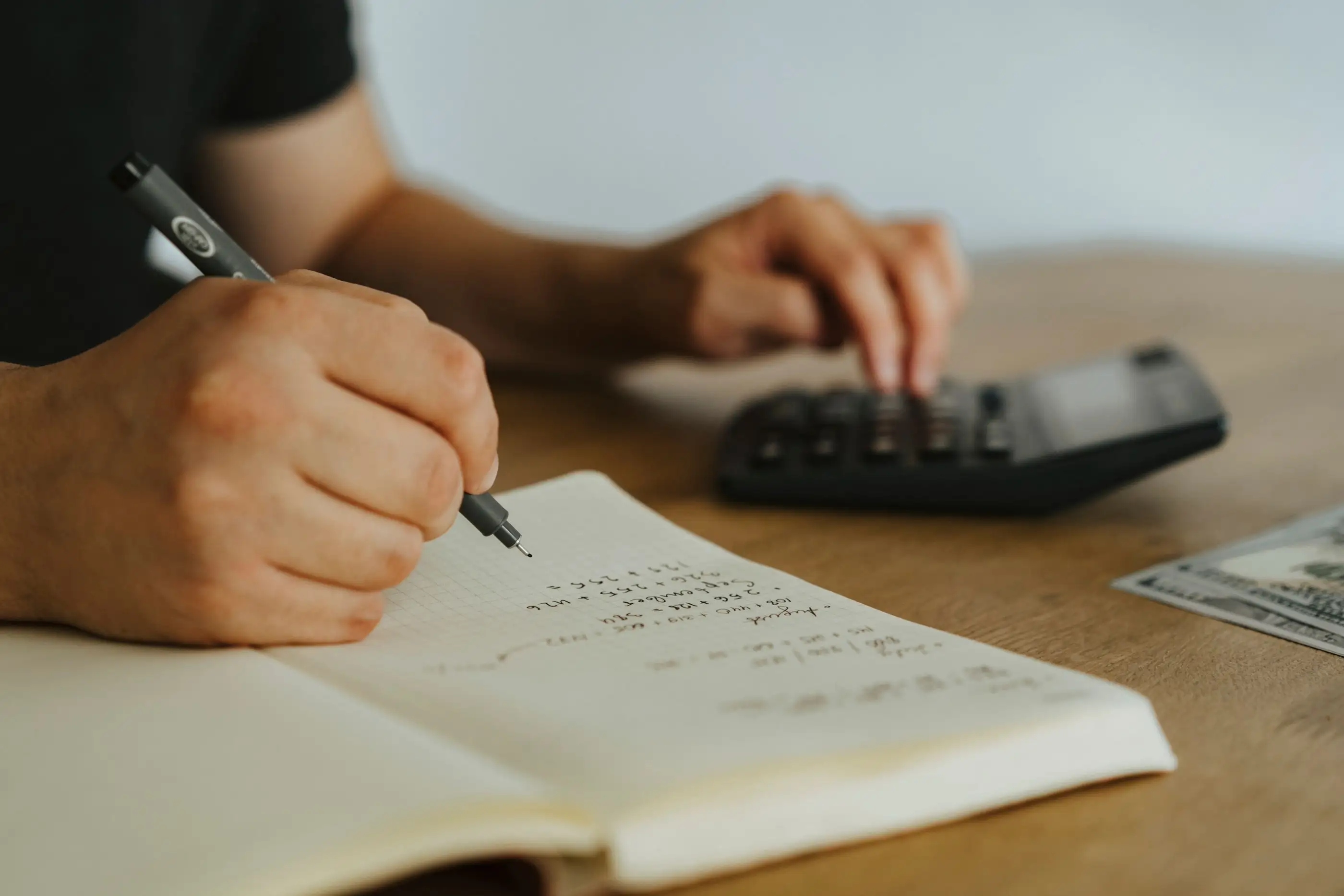 A hand writes figures in a notebook beside a calculator.