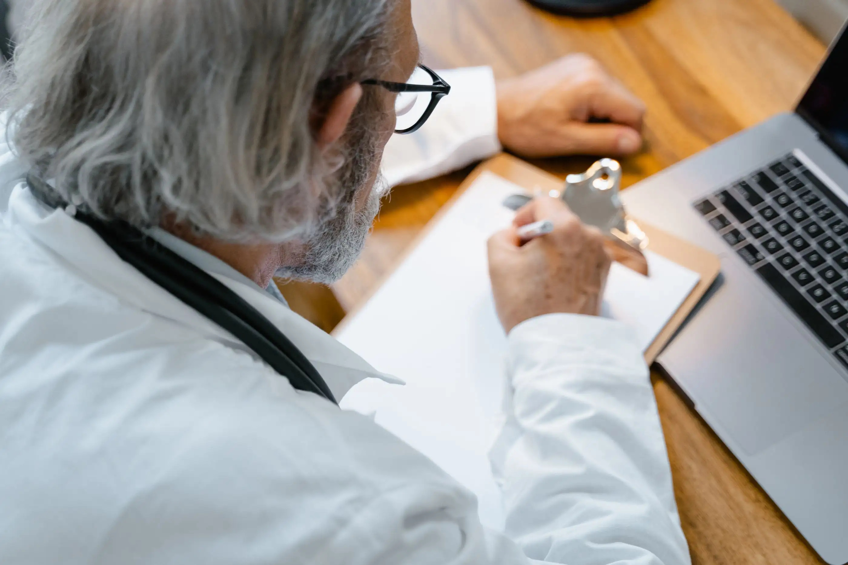 An older physician writes notes beside his laptop.
