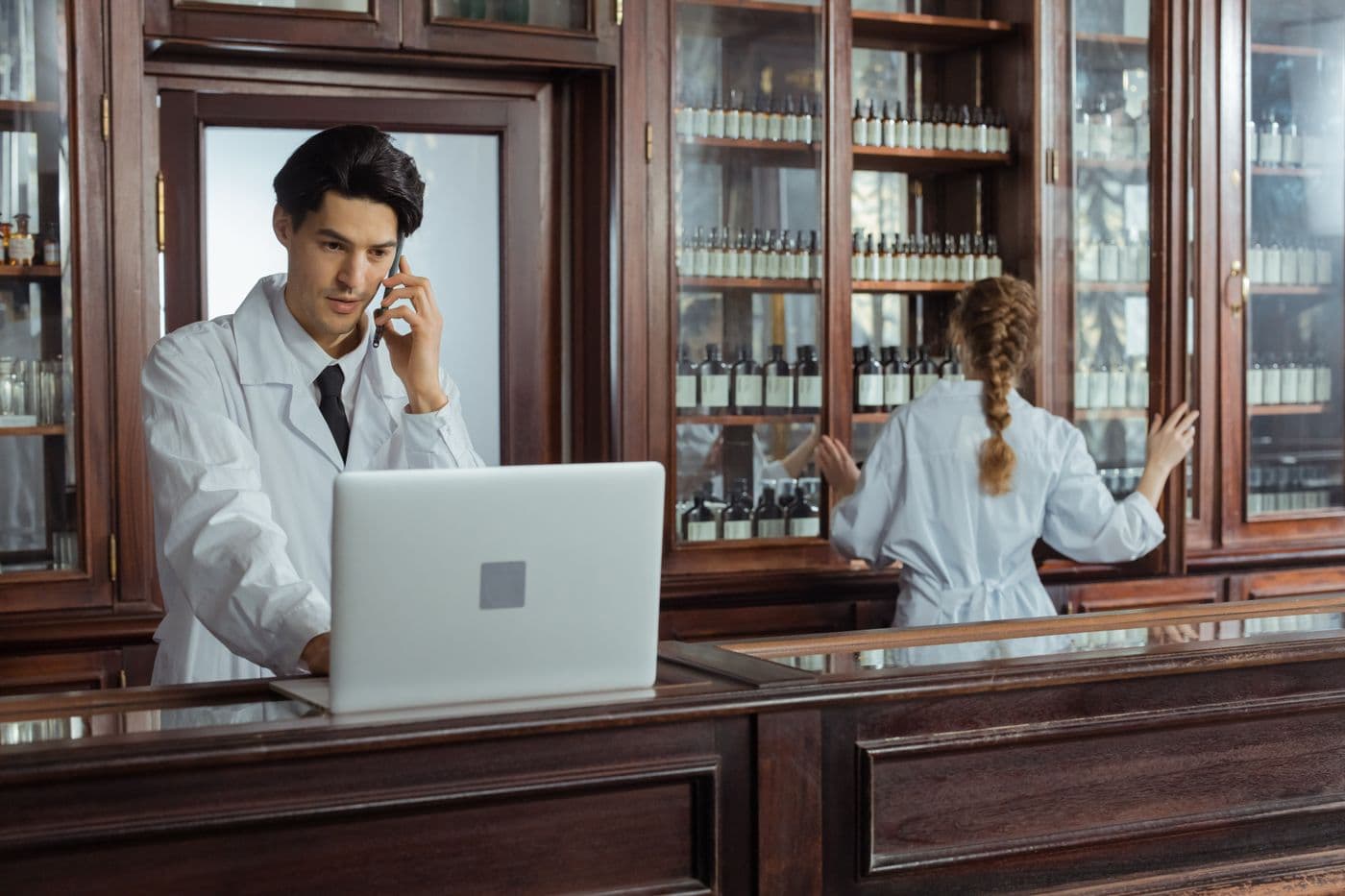 A pharmacist talks on the phone at a laptop while a colleague works at the shelves behind him.
