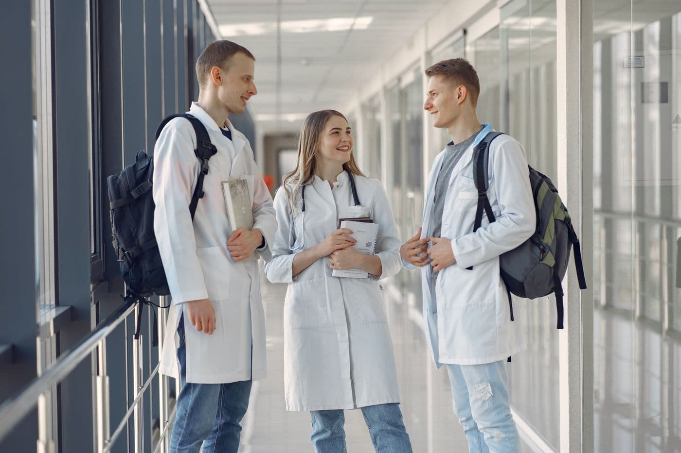Three young clinicians in white coats talk in a hospital corridor.