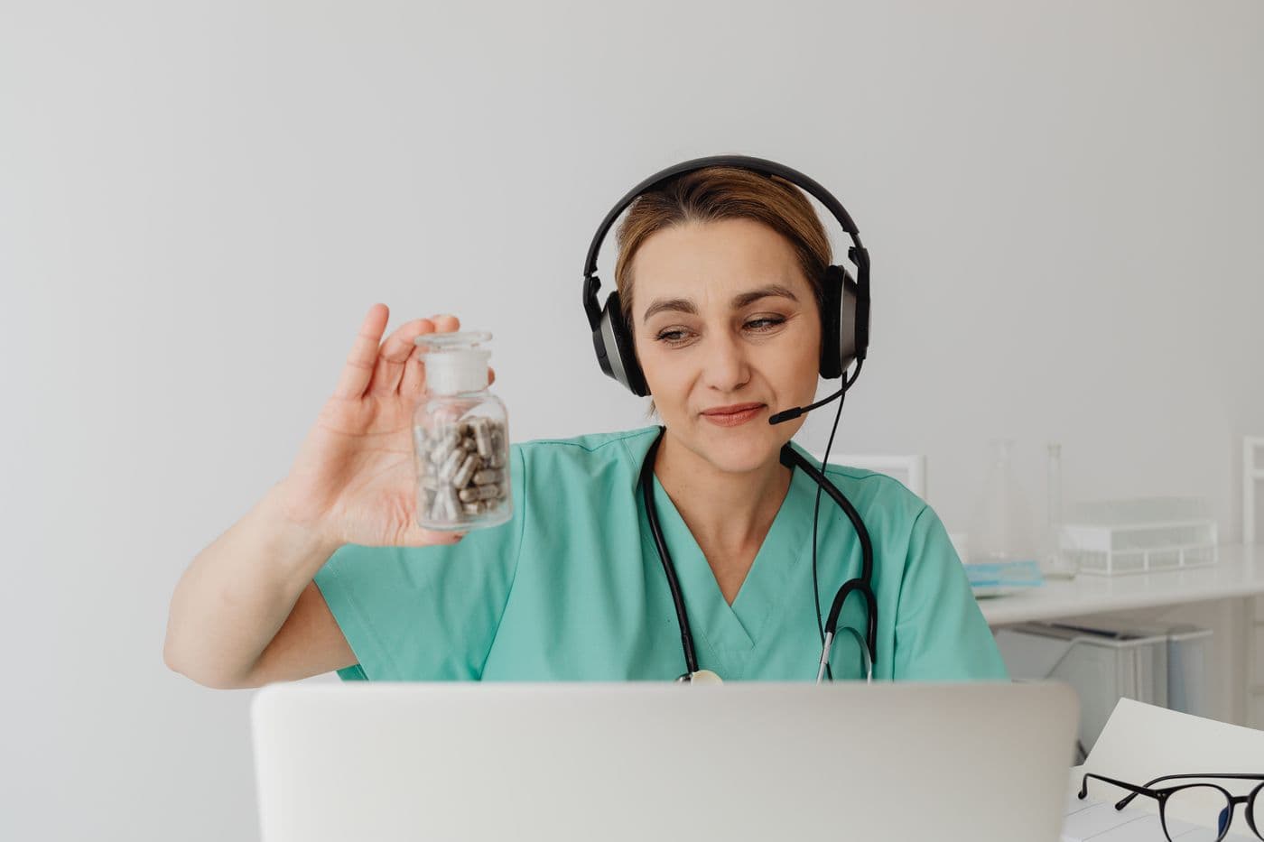 A clinician wearing a headset holds up a bottle of tablets during a video consultation.
