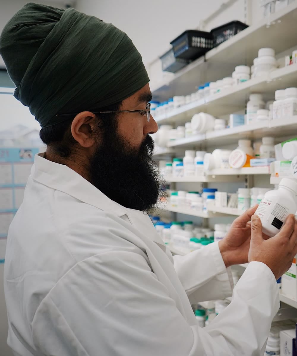 A pharmacist in a white coat checks the label on a medicine bottle in front of well-stocked pharmacy shelves.
