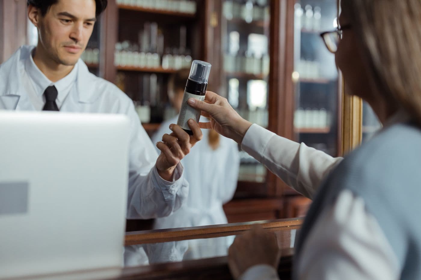 A pharmacist in a white coat hands a medicine bottle to a patient across the pharmacy counter.