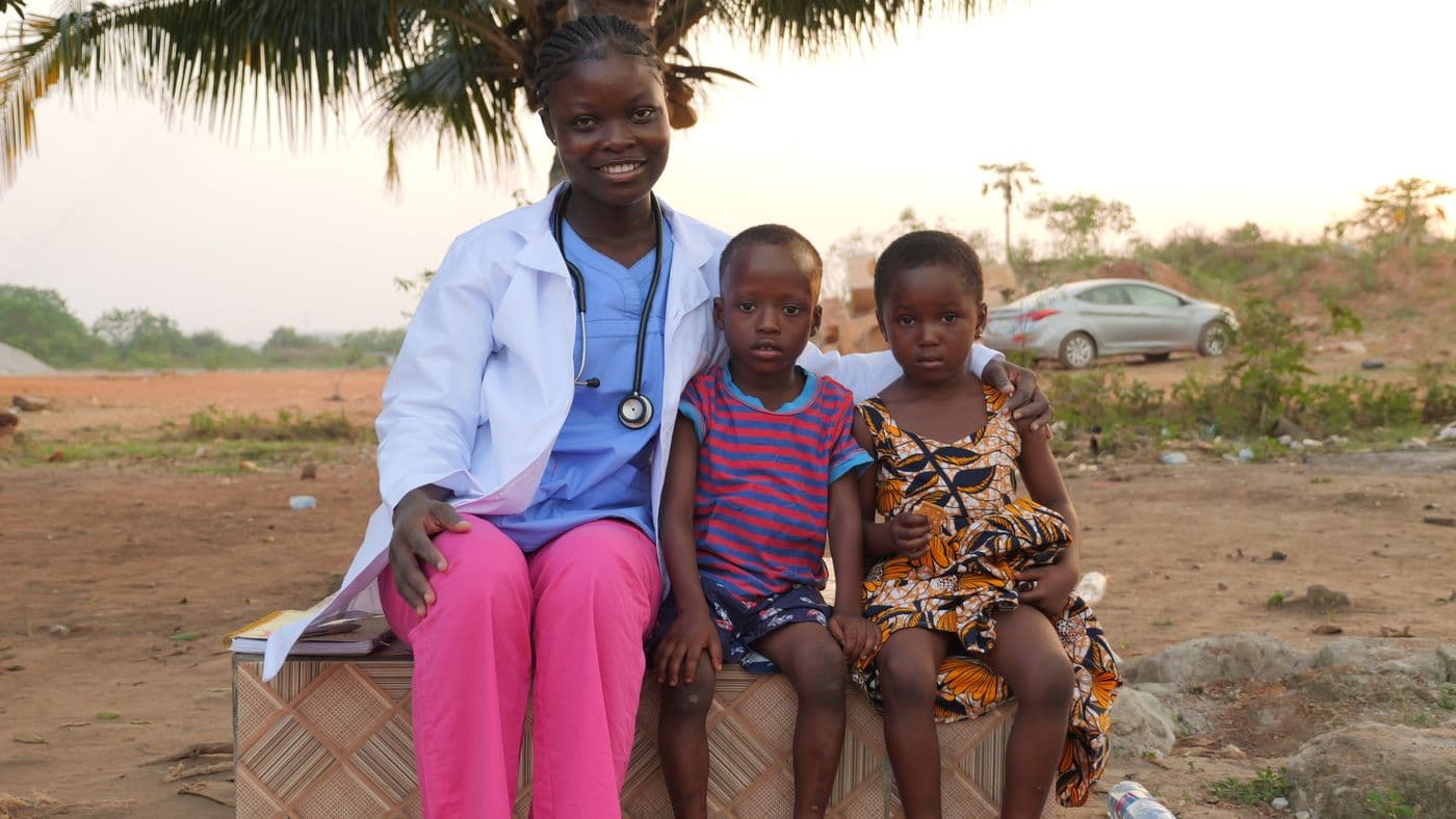 A community health worker sits outdoors with two young children.
