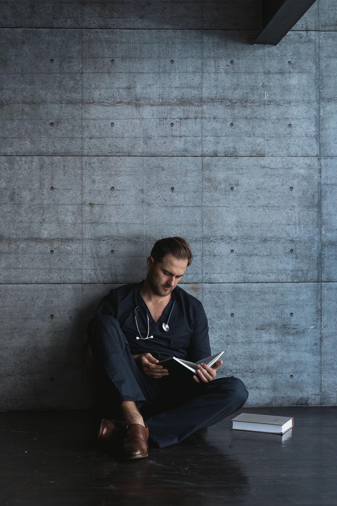 A clinician in scrubs sits on the floor against a concrete wall, reading.
