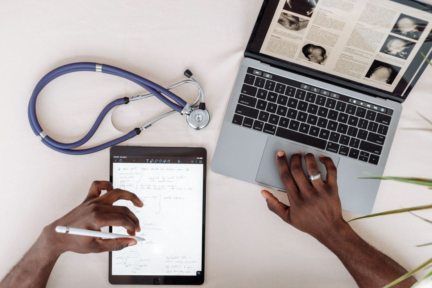 A clinician takes notes on a tablet beside a laptop and a stethoscope.