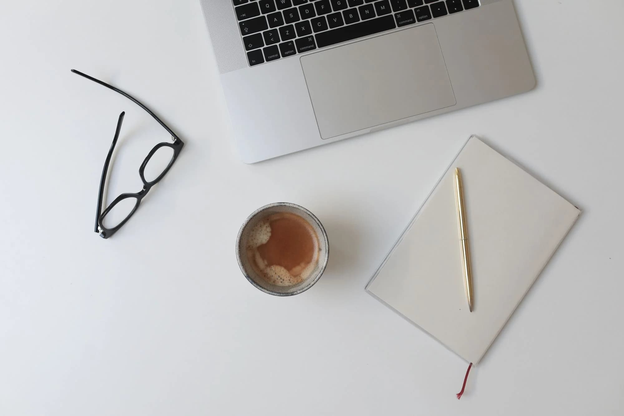 A notebook, laptop, glasses and coffee arranged on a desk.