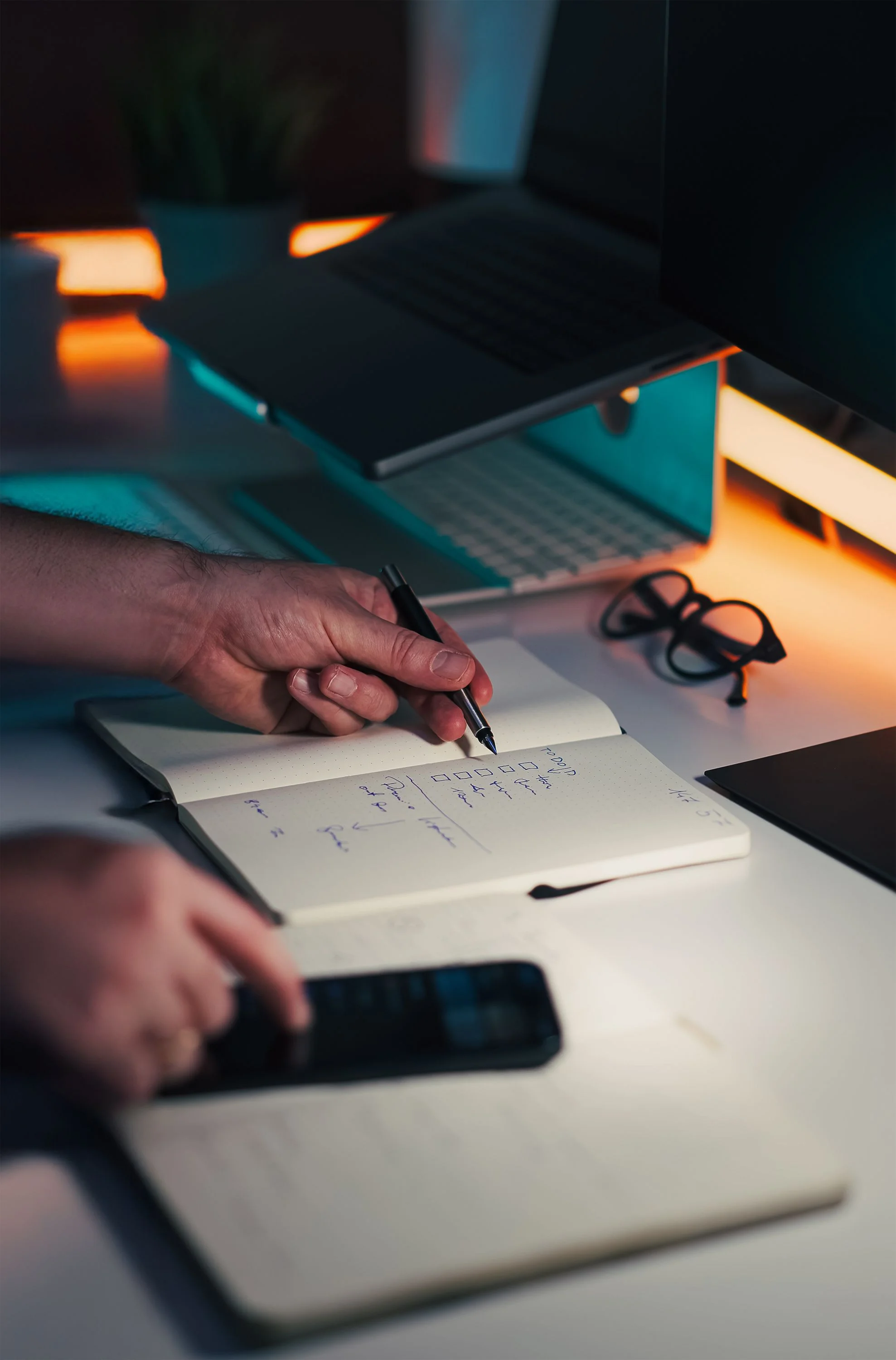 Hands make notes at a desk with a laptop and phone.