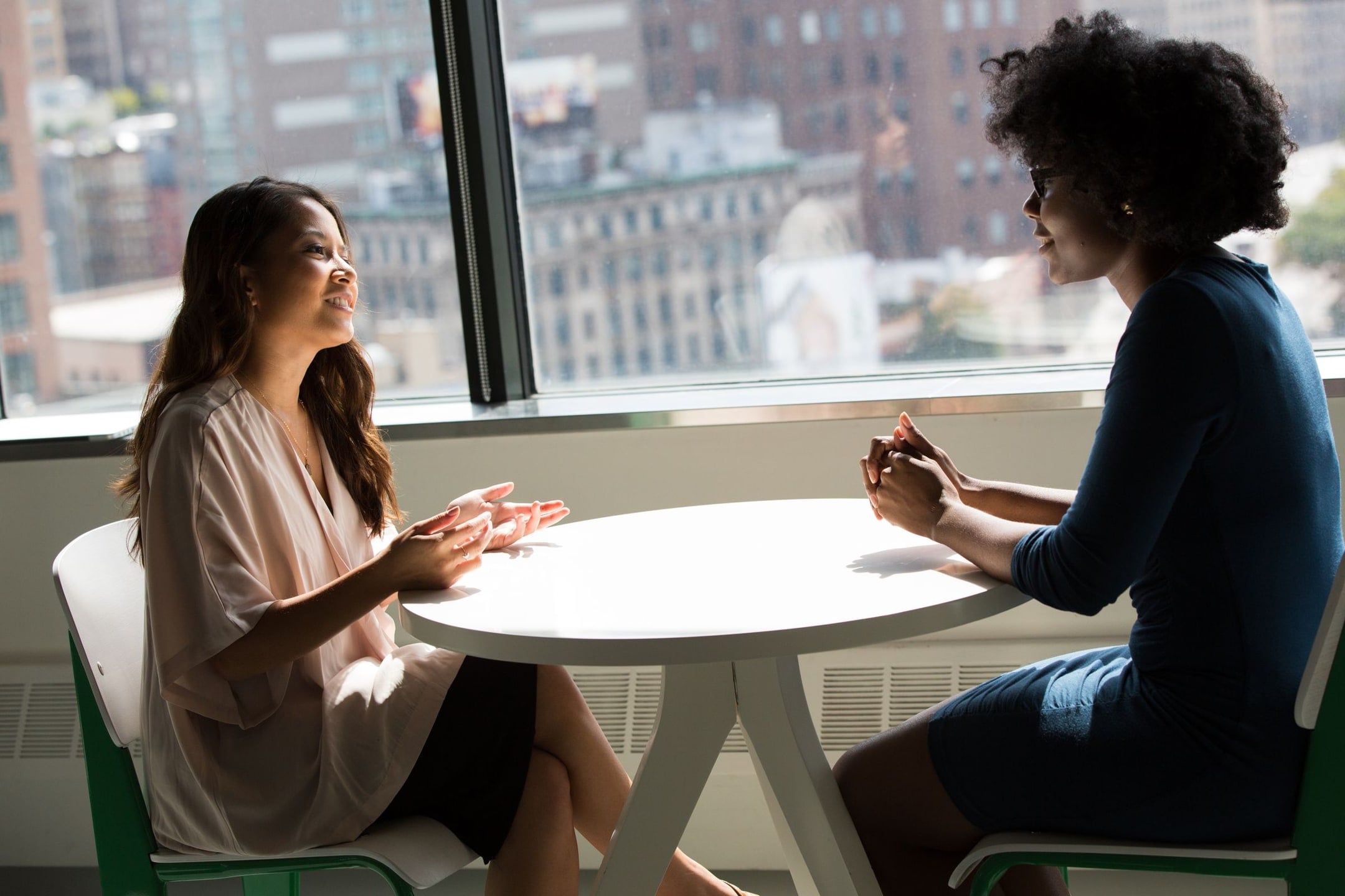 Two professionals talk across a table by a bright office window.