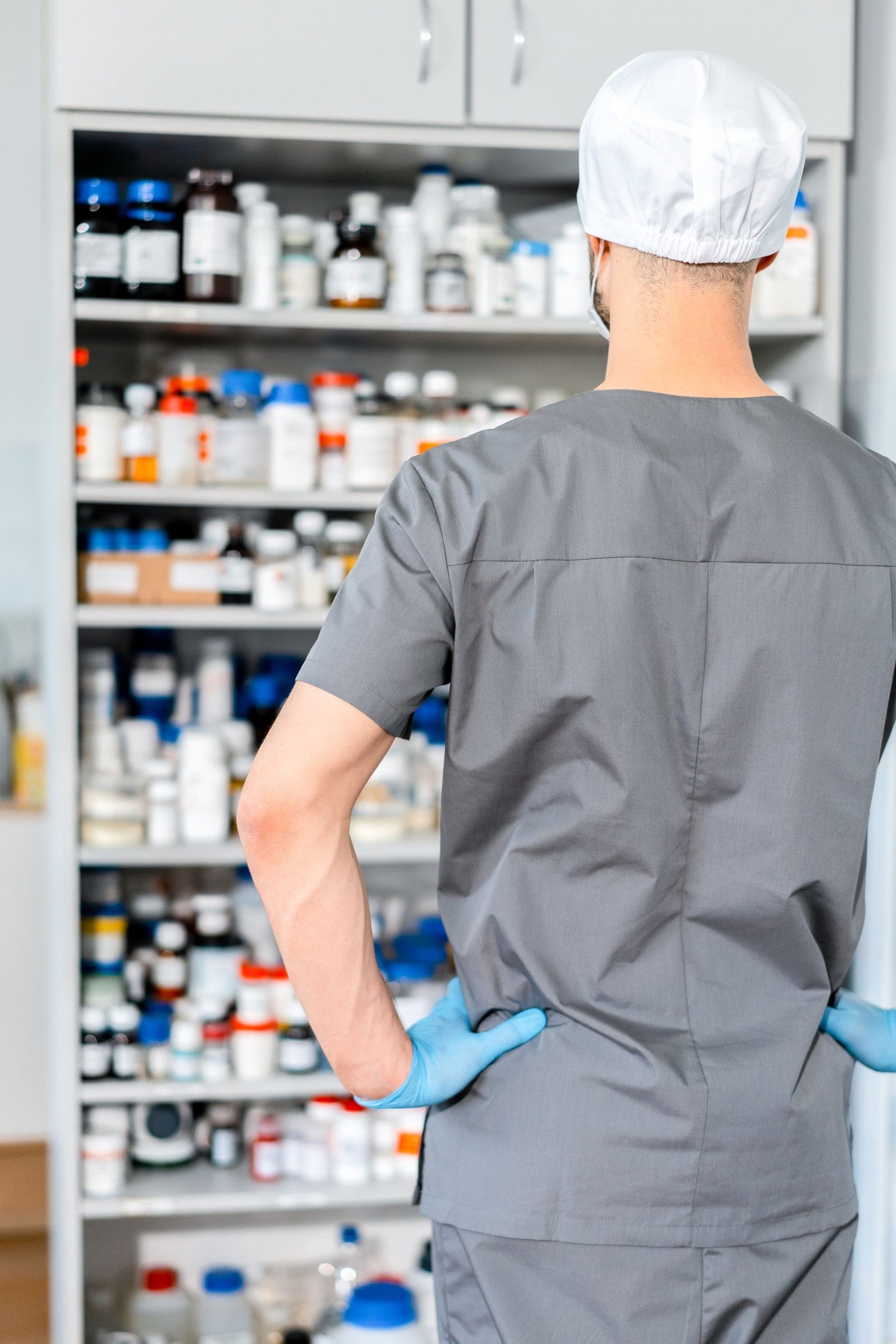 A pharmacist in scrubs and gloves at shelves stocked with medications.